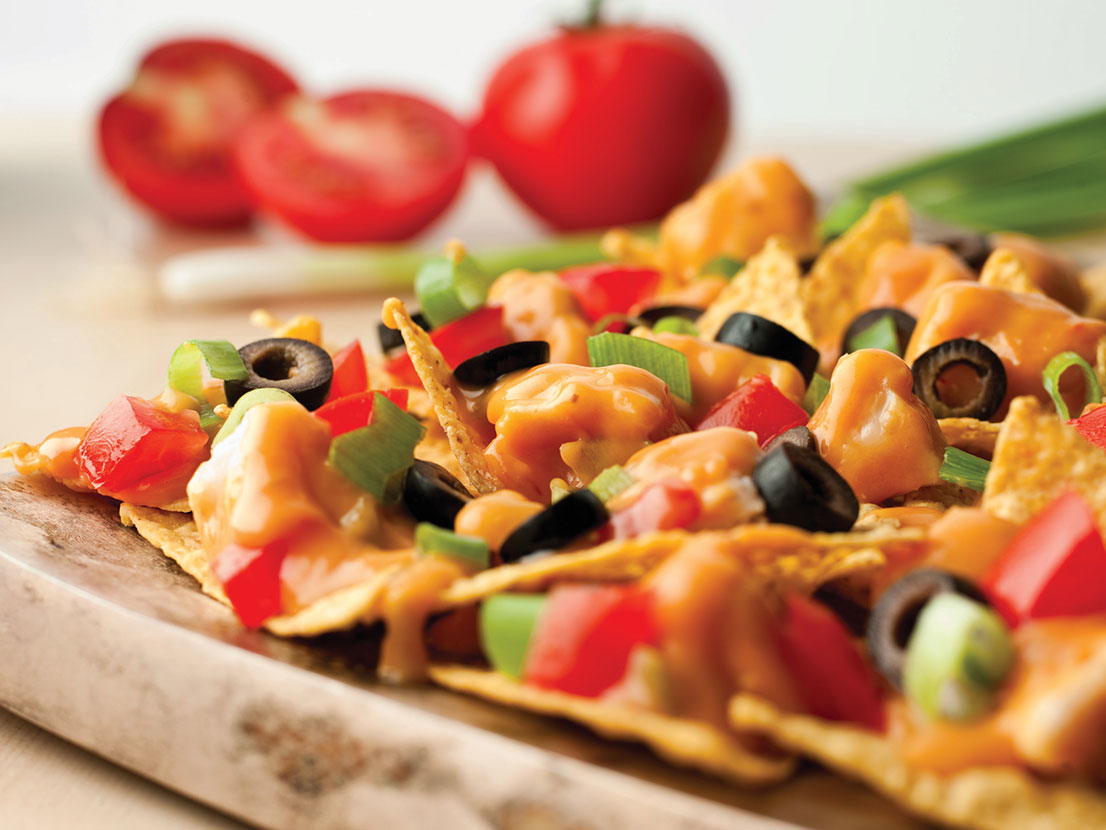 An image of prepared Chicken Nachos made with Pace® Chunky Salsa, Campbell's Condensed Cheddar Cheese Soup, Swanson® Premium White Chunk Chicken, tortilla chips, tomato, green onion and black olives.