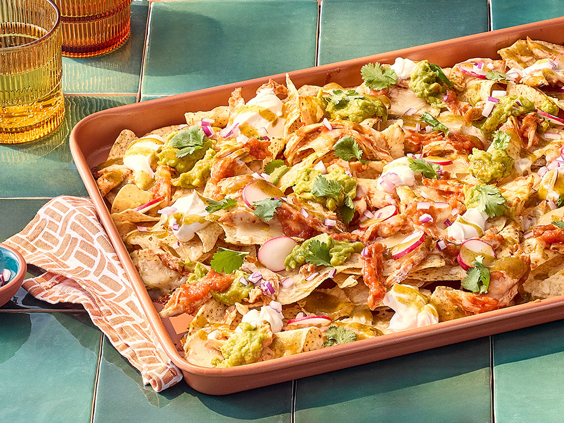 An image of prepared Chicken Tinga Nachos made with Pace® Chunky Salsa, Pace® Signature Sauces Hot Honey Chipotle Sauce, chicken, tortilla chips, Monterey Jack cheese, sour cream, guacamole, Pace® Signature Sauces Cilantro Jalapeño Lime Sauce, radishes and cilantro.
