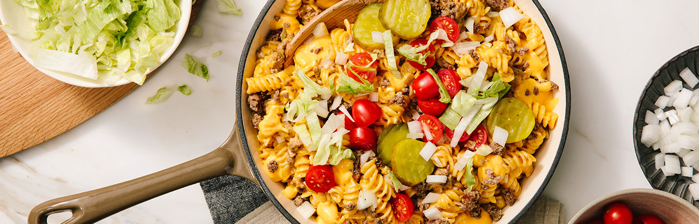 An image of prepared Bacon Cheeseburger Pasta Skillet made with Prego® Creamy Cheddar Sauce, ground beef, rotini, onion, dill pickles, cherry tomatoes and lettuce.