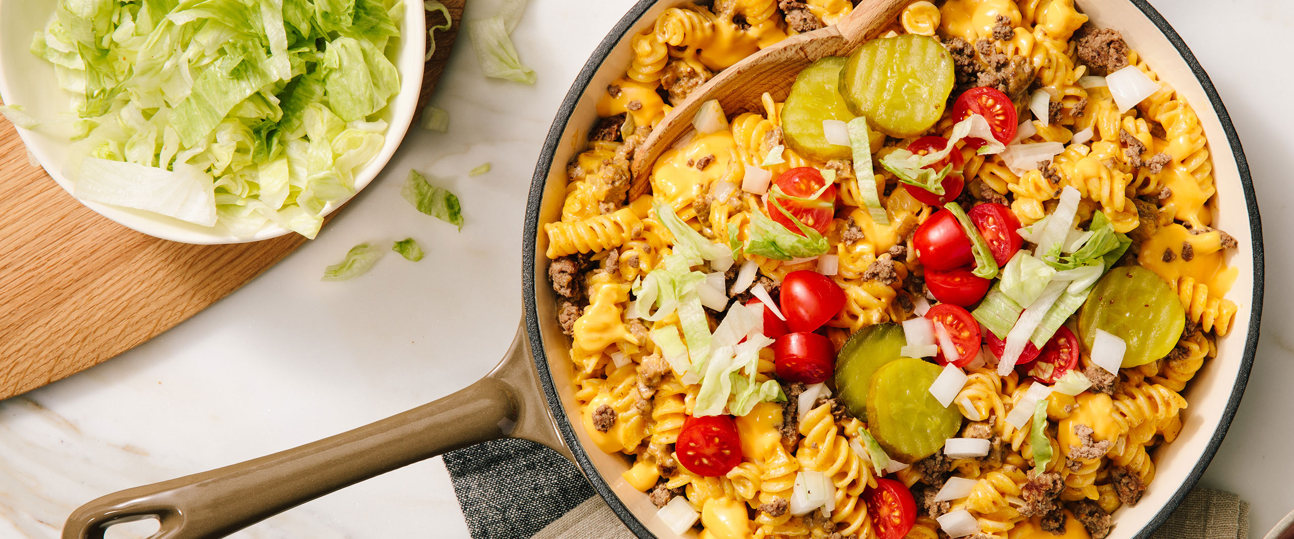An image of prepared Bacon Cheeseburger Pasta Skillet made with Prego® Creamy Cheddar Sauce, ground beef, rotini, onion, dill pickles, cherry tomatoes and lettuce.