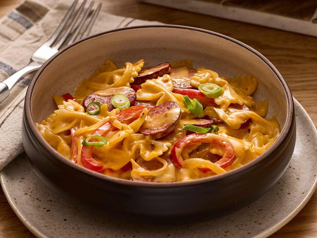 An image of prepared One Pot Smoky Cheddar Kielbasa Pasta made with Prego® Cheesy Bacon Cheddar Cheese Sauce, turkey kielbasa, onion, bell pepper, farfalle and green onion.