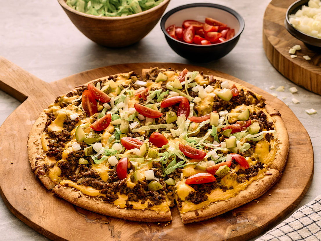 An image of prepared Bacon Cheeseburger Pizza made with ground beef, onion, Prego® Cheesy Bacon Cheddar Cheese Sauce, Cheddar cheese, dill pickle and cherry tomatoes.