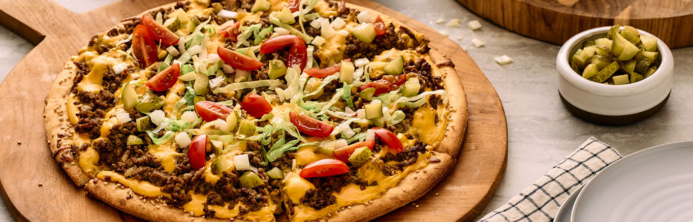 An image of prepared Bacon Cheeseburger Pizza made with ground beef, onion, Prego® Cheesy Bacon Cheddar Cheese Sauce, Cheddar cheese, dill pickle and cherry tomatoes.
