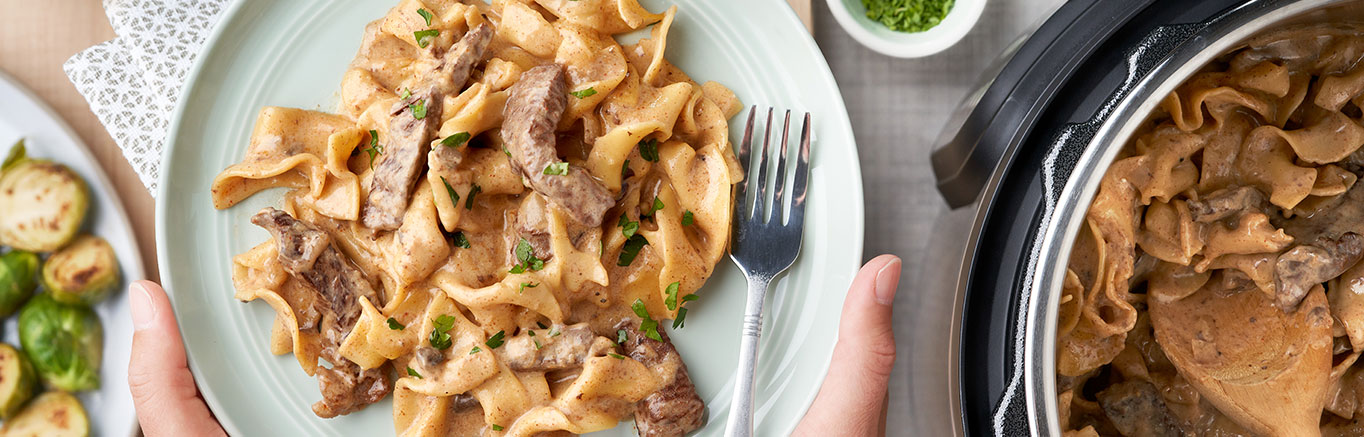 An image of prepared Instant Pot® Easy Beef Stroganoff showing onions, beef sirloin steak, Swanson® Beef Broth, Campbell's® Condensed Cream of Mushroom Soup, sour cream, parsley and egg noodles.