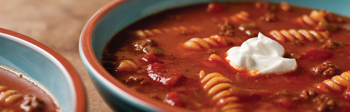 An image of prepared Taco Twist Soup made with ground beef, chili powder, Swanson® Beef Broth, Pace® Picante Sauce, canned diced tomatoes, rotini pasta and sour cream.