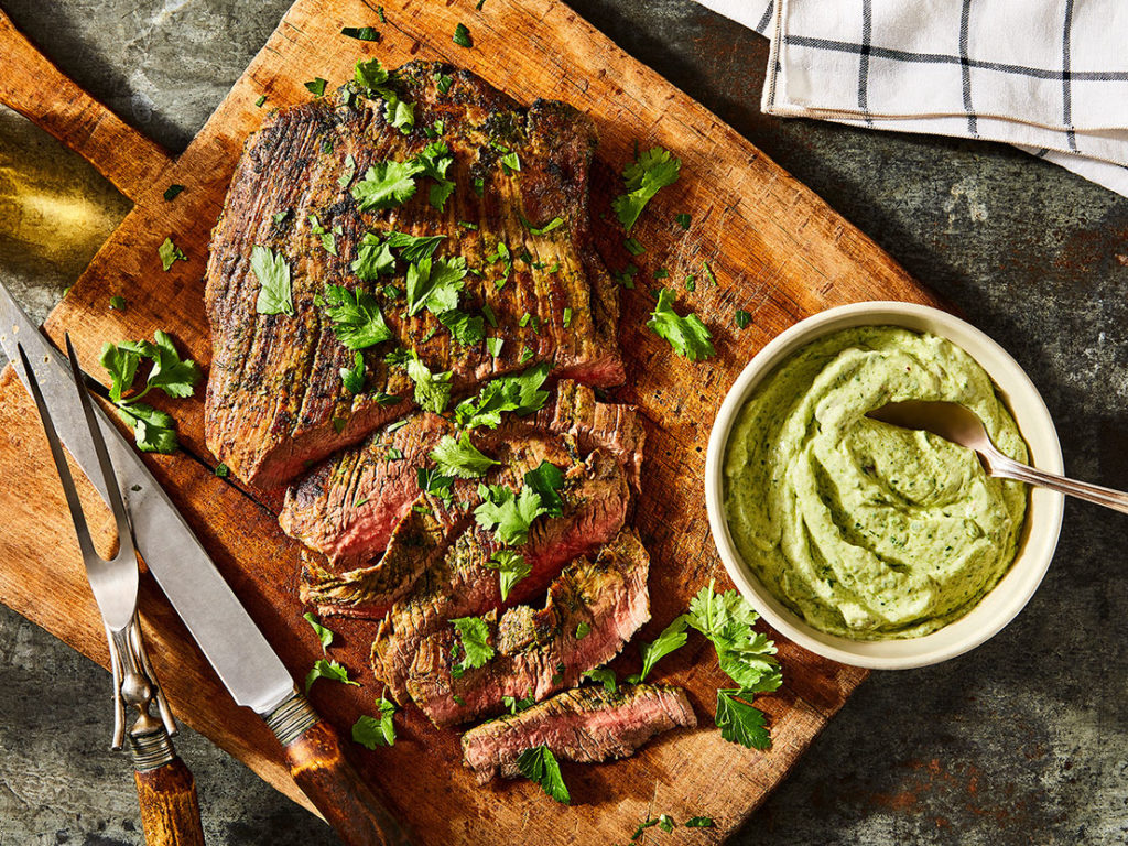 An image of prepared Big Batch Flank Steak with Creamy Chimichurri Sauce showing beef flank steak with parsley, cilantro and garlic on top and Campbell's® Condensed Unsalted Cream of Mushroom Soup with sour cream on the side.