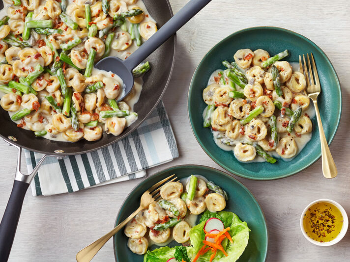 An image of prepared Creamy Tortellini with Asparagus in Mushroom Bacon Sauce showing Campbell's® Condensed Cream of Cremini & Shiitake Soup, Swanson® Natural Goodness® Chicken Broth, asparagus, cheese tortellini and bacon.